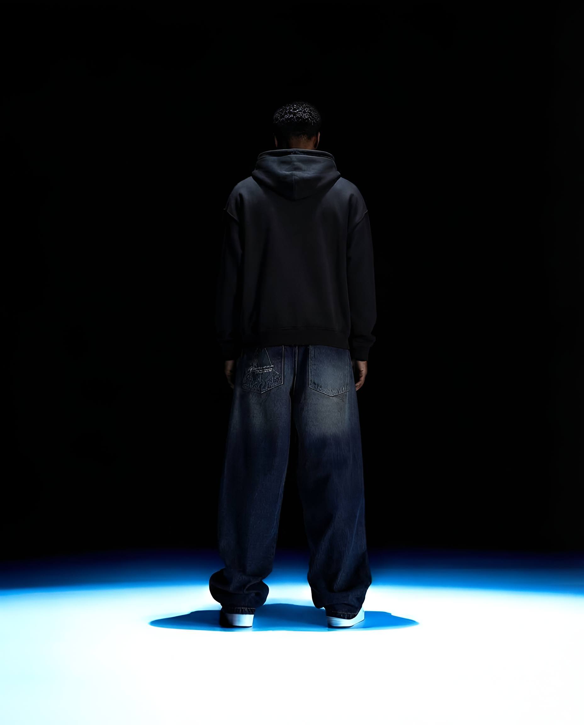Person wearing a dark hoodie and jeans standing in a dark room with blue light on the floor.